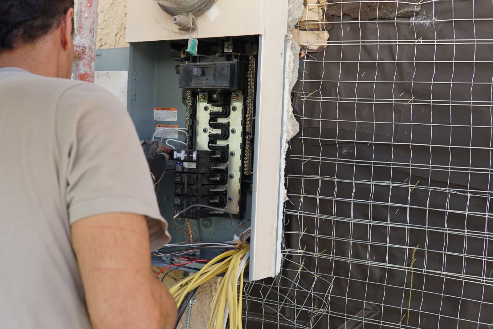 An electrician working on an open electrical panel, surrounded by exposed wires and construction material, performing maintenance or installation work.