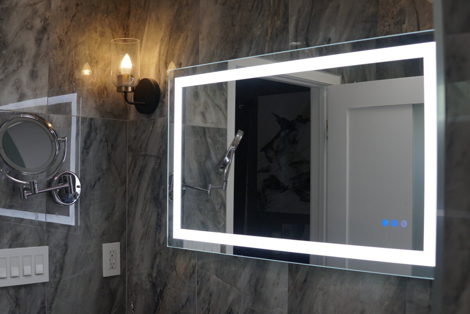 A modern bathroom featuring a lighted mirror with integrated LED lighting, a wall-mounted magnifying mirror, and a sconce light fixture against marble wall tiles.